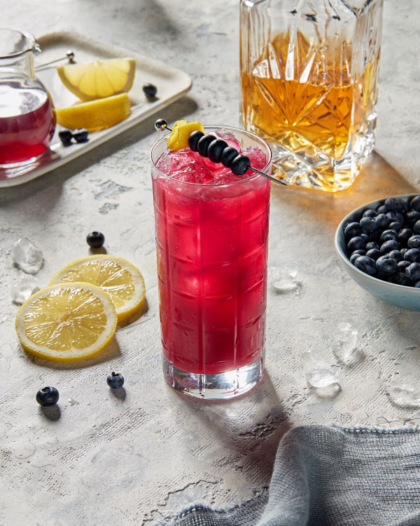 Un verre highball de fizz au whisky aux bleuets sur glace, garni d'une brochette de bleuets, présenté avec une carafe de whisky, une carafe de sirop simple, des quartiers et rondelles de citron, ainsi qu'un bol de bleuets.