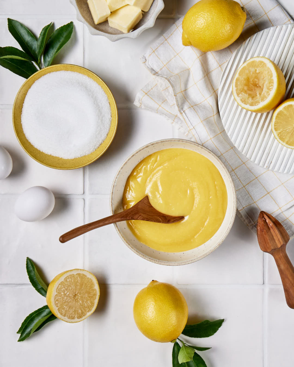 Curd au citron Un bol de lemon curd est présenté sur un comptoir carrelé, avec des citrons entiers et coupés en deux, un presse-citron et un bol de sucre granulé.
