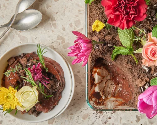 Pouding au chocolat saupoudré de miettes de biscuits et garni de fleurs comestibles dans un plat à four en verre, présenté avec une portion de pudding dans un bol et des cuillères.