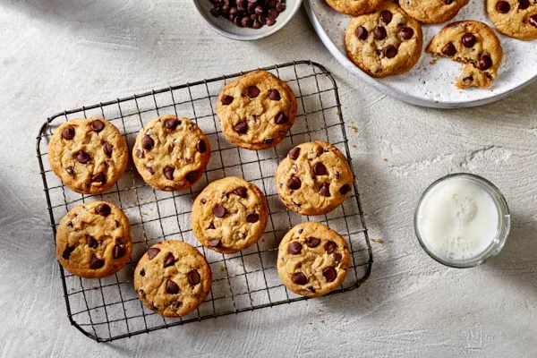 Des biscuits aux pépites de chocolat sur une grille de refroidissement en fil métallique, avec des biscuits supplémentaires sur un plateau, montrés avec un bol de pépites de chocolat et un verre de lait blanc. 