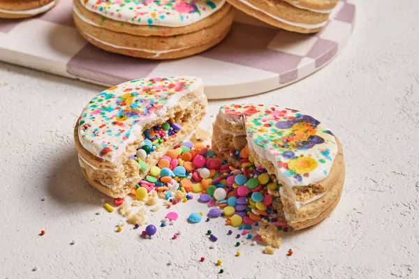 Un biscuit en forme de piñata glacé, brisé en deux avec des confettis are-en-ciel et des vermicelles sortis, présenté avec trois autres biscuits sur un plateau à carreaux blancs et mauves en arrière-plan