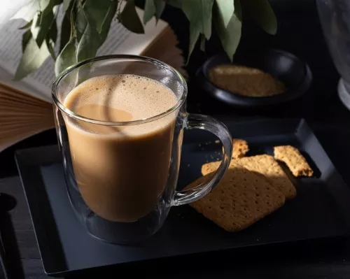Tasse en verre remplie de kopi cham sur un plateau avec des biscuits, un livre et une plante