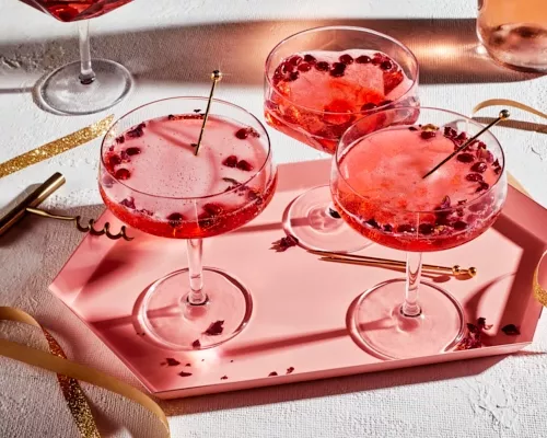Trois cocktails pétillants à la grenade et à la rose dans des verres sur un plateau rose.