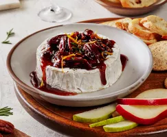  Brie cuit au four, garni de noix de pécan confites épicées et de sirop de Cassonade Foncée et décoré de zeste d'orange et de romarin, présenté sur une assiette sur un plateau de service avec des craquelins, des crostinis et des tranches de poires et de pommes.