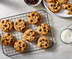 Des biscuits aux pépites de chocolat sur une grille de refroidissement en fil métallique, avec des biscuits supplémentaires sur un plateau, montrés avec un bol de pépites de chocolat et un verre de lait blanc. 