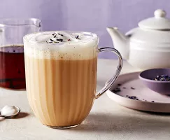 Un verre à latte London Fog avec mousse de lait, montré avec une carafe de sirop de vanille, une théière, une cuillère à mélanger et un bol pourpre de thé Earl Grey en vrac sur un plateau.