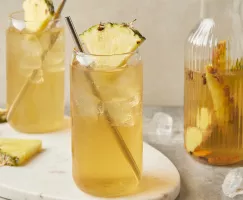 Deux verres de tepache avec des pailles en métal et garnis de quartiers d'ananas, présentés avec une carafe de tepache.