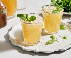 Deux verres de julep à la menthe sans alcool avec de la glace pilée et garnis de menthe, servis sur un plateau blanc et présentés avec un bol de feuilles de menthe, une moitié de lime, une carafe de julep préparé et une carafe de sirop simple à la menthe.