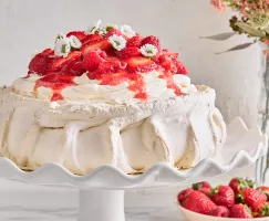 Une Pavlova garnie de framboises, de fraises et de fleurs sur un support à gâteau blanc, présentée avec un vase de fleurs et un bol de baies.