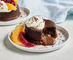 Deux gâteaux individuels au chocolat fondant garnis de crème fouettée et de cacao en poudre, agrémentés de segments d'orange et d'orange sanguine. L'un des gâteaux est montré avec un morceau manquant, laissant échapper une sauce au chocolat à partir du centre
