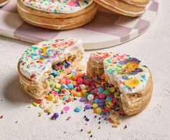 Un biscuit en forme de piñata glacé, brisé en deux avec des confettis are-en-ciel et des vermicelles sortis, présenté avec trois autres biscuits sur un plateau à carreaux blancs et mauves en arrière-plan