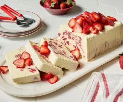 Un pain rectangulaire de semifreddo à la fraise et à la fleur de sureau sur un plateau en marbre, garni de tranches de fraises, avec trois tranches coupées, présenté avec trois assiettes blanches à bordure rouge, trois cuillères rouges et un bol rouge de fraises.