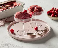 Deux coupes à pied de sorbet aux cerises garnies de cerises, sur un plateau rose pâle avec des cuillères en or, présentées avec un bol de cerises et une casserole de sorbet aux cerises avec une cuillère à boule pour servir.