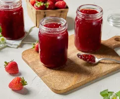 Trois pots de confiture de fraises, deux sur une planche à découper en bois et un sur un torchon, présentés avec une cuillère de confiture, des fraises fraîches et des fleurs de fraises.