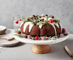 Un gâteau Bundt au pain d’épices glacé déposé sur une assiette sur pied et décoré de branches de romarin et de canneberges confi