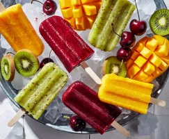 Une variété de paletas de fruits congelés dans un seau de glace, servis avec des kiwis, des cerises et des mangues.