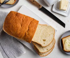Une miche partiellement tranchée sur une planche à découper avec un couteau à pain et une tranche beurrée sur une assiette
